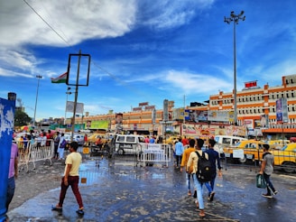 A bustling city scene with people walking on a wet street, surrounded by vibrant yellow taxis and vehicles. Buildings in the background display various advertisements and signs. The sky is a bright blue with scattered clouds, and a large Indian flag is mounted on a tall pole.