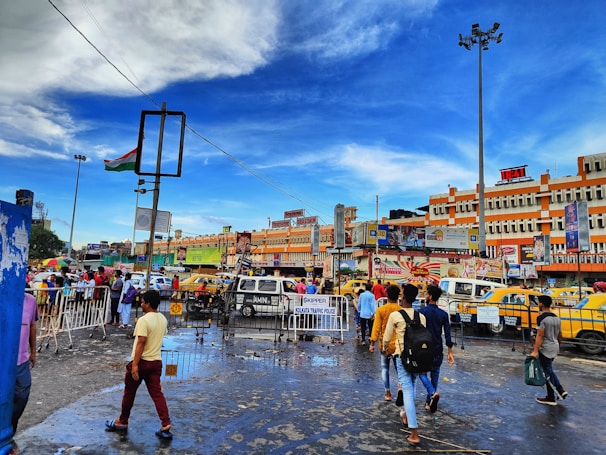 A bustling city scene with people walking on a wet street, surrounded by vibrant yellow taxis and vehicles. Buildings in the background display various advertisements and signs. The sky is a bright blue with scattered clouds, and a large Indian flag is mounted on a tall pole.