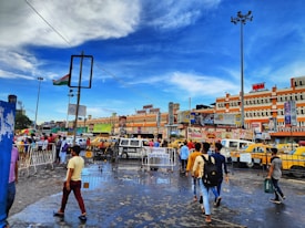 A bustling city scene with people walking on a wet street, surrounded by vibrant yellow taxis and vehicles. Buildings in the background display various advertisements and signs. The sky is a bright blue with scattered clouds, and a large Indian flag is mounted on a tall pole.