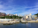 A coastal urban landscape featuring a calm body of water in the foreground, with a sandy beach lined with rocks and a few tents. Behind the beach, there is a bustling cityscape with high-rise buildings and greenery. The sky is clear and blue, contributing to a vibrant, lively scene.