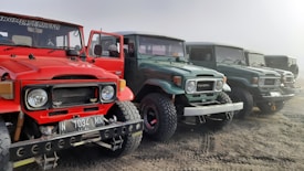 A row of vintage Toyota Land Cruiser vehicles is parked on a sandy terrain. The front vehicle is bright red, followed by various shades of green and gray ones. The cars have rugged tires, and the red one has an open driver's side door.