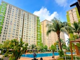 Tall apartment buildings with light green and beige exteriors rise behind a courtyard featuring a clear, inviting swimming pool surrounded by lush palm trees and other greenery. A bright blue sky with scattered clouds provides a backdrop.