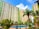Tall apartment buildings with light green and beige exteriors rise behind a courtyard featuring a clear, inviting swimming pool surrounded by lush palm trees and other greenery. A bright blue sky with scattered clouds provides a backdrop.