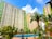 Tall apartment buildings with light green and beige exteriors rise behind a courtyard featuring a clear, inviting swimming pool surrounded by lush palm trees and other greenery. A bright blue sky with scattered clouds provides a backdrop.