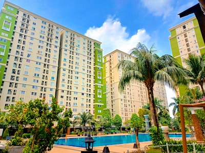 Tall apartment buildings with light green and beige exteriors rise behind a courtyard featuring a clear, inviting swimming pool surrounded by lush palm trees and other greenery. A bright blue sky with scattered clouds provides a backdrop.