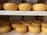 Several round, golden-yellow cheese wheels are neatly arranged on two wooden shelves. The cheese wheels appear smooth and have a glossy finish, indicating freshness or a wax coating. The background suggests an environment typical for cheese storage or aging.