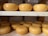 Several round, golden-yellow cheese wheels are neatly arranged on two wooden shelves. The cheese wheels appear smooth and have a glossy finish, indicating freshness or a wax coating. The background suggests an environment typical for cheese storage or aging.