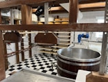 A room equipped for cheesemaking with wooden frames holding metal presses, a large circular stainless steel vat, and shelves storing several wheels of cheese. The space features a black-and-white checkered tile floor and white tile walls. Overhead wooden beams and various tools and equipment are also visible.