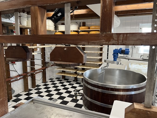 A room equipped for cheesemaking with wooden frames holding metal presses, a large circular stainless steel vat, and shelves storing several wheels of cheese. The space features a black-and-white checkered tile floor and white tile walls. Overhead wooden beams and various tools and equipment are also visible.