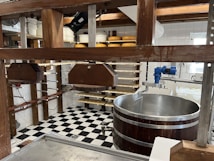 A room equipped for cheesemaking with wooden frames holding metal presses, a large circular stainless steel vat, and shelves storing several wheels of cheese. The space features a black-and-white checkered tile floor and white tile walls. Overhead wooden beams and various tools and equipment are also visible.