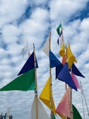 A collection of colorful triangular flags attached to wooden poles is set against a backdrop of a cloudy sky. The flags feature a variety of colors, including blue, green, yellow, pink, and white, and are spaced at irregular intervals, creating an eye-catching display.