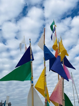 A collection of colorful triangular flags attached to wooden poles is set against a backdrop of a cloudy sky. The flags feature a variety of colors, including blue, green, yellow, pink, and white, and are spaced at irregular intervals, creating an eye-catching display.