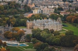 An aerial view of an expansive historic palace surrounded by lush green gardens and trees with autumnal hues. The palace features a traditional architectural style with a prominent stone facade and multiple chimneys. In the background, a densely built urban landscape with residential buildings and roads can be seen.