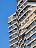 A modern high-rise building with an angular design is under construction. The facade features multiple balconies with blue glass railings. The structure casts geometric shadows against a clear blue sky, with some scaffolding visible on the right side.
