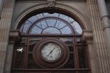An ornate architectural facade features a semi-circular window above a large round barometer. The structure is framed by stone columns with decorative elements, and the barometer is surrounded by inscriptions indicating weather conditions.
