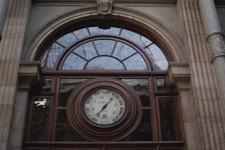 An ornate architectural facade features a semi-circular window above a large round barometer. The structure is framed by stone columns with decorative elements, and the barometer is surrounded by inscriptions indicating weather conditions.