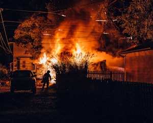 A large fire is engulfing a building, with intense flames and thick smoke illuminating the night sky. A silhouette of a person appears to be approaching the fire, possibly a firefighter. Nearby structures and trees are partially visible, and power lines cross the scene above the flames.