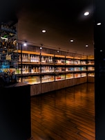 A dimly lit upscale interior of a liquor store or bar featuring a wide selection of illuminated bottles arranged on wooden shelves. The floor is made of dark polished wood, and the ceiling has several recessed lights providing focused illumination.