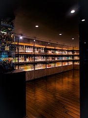 A dimly lit upscale interior of a liquor store or bar featuring a wide selection of illuminated bottles arranged on wooden shelves. The floor is made of dark polished wood, and the ceiling has several recessed lights providing focused illumination.