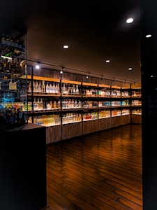 A dimly lit upscale interior of a liquor store or bar featuring a wide selection of illuminated bottles arranged on wooden shelves. The floor is made of dark polished wood, and the ceiling has several recessed lights providing focused illumination.