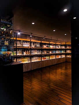 A dimly lit upscale interior of a liquor store or bar featuring a wide selection of illuminated bottles arranged on wooden shelves. The floor is made of dark polished wood, and the ceiling has several recessed lights providing focused illumination.