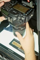 A digital camera is being held, focused on a film negative placed on a lightbox. The camera's screen displays an image with film sprockets visible. The hand appears to be adjusting the zoom or focus. The brand name Canon is visible on the camera, and a Gaomon lightbox is used to illuminate the film.