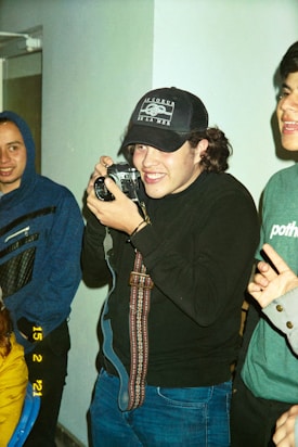 A group of young adults, one of whom is holding a vintage camera and smiling, while another person stands in the background wearing a hoodie. The setting appears casual and indoors, with a sense of camaraderie.