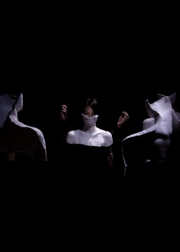 A person partially concealed by a white cloth appears surrounded by three white plaster busts in a dark setting. The scene is dramatic, with a focus on contrast between light and shadow, highlighting the person's eyes and hands that emerge from the darkness. The busts are positioned symmetrically, adding a sense of mystery and surrealism.
