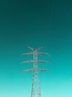 A tall metal electricity pylon stands prominently against a vast, clear turquoise sky. Numerous electrical wires extend symmetrically from the top of the pylon, creating a sense of order and structure.