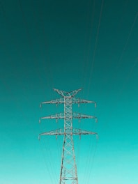 A tall metal electricity pylon stands prominently against a vast, clear turquoise sky. Numerous electrical wires extend symmetrically from the top of the pylon, creating a sense of order and structure.