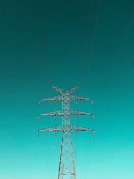 A tall metal electricity pylon stands prominently against a vast, clear turquoise sky. Numerous electrical wires extend symmetrically from the top of the pylon, creating a sense of order and structure.
