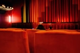 A movie theater interior featuring rows of red upholstered seats facing a large stage with a closed red curtain. Soft lighting illuminates the walls, creating a warm and cozy ambiance. Only a few seats are occupied, contributing to a sense of spaciousness.