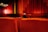 A movie theater interior featuring rows of red upholstered seats facing a large stage with a closed red curtain. Soft lighting illuminates the walls, creating a warm and cozy ambiance. Only a few seats are occupied, contributing to a sense of spaciousness.