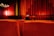 A movie theater interior featuring rows of red upholstered seats facing a large stage with a closed red curtain. Soft lighting illuminates the walls, creating a warm and cozy ambiance. Only a few seats are occupied, contributing to a sense of spaciousness.