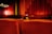 A dimly lit theater room featuring red velvet seats arranged in rows. The curtains in the background are also red, with a soft glow from ambient lighting. A person with glasses is partially visible, peeking over the seats.