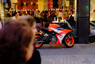 A brightly colored, sleek motorcycle is parked on the sidewalk outside a busy café or restaurant. Inside, several people are seated at tables, engaged in conversation and dining. The scene is set in the evening, illuminated by artificial lights from the interior and streetlights. The motorcycle features a vivid orange and black design, and the people inside are casually dressed, appearing relaxed and social.