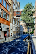 A narrow urban street is lined with brick buildings, one of which has a sign reading 'London Metropolitan University'. The street features parked bicycles, a few pedestrians, and a tree casting shadows across the road. The sky is clear and blue.