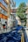 A narrow urban street is lined with brick buildings, one of which has a sign reading 'London Metropolitan University'. The street features parked bicycles, a few pedestrians, and a tree casting shadows across the road. The sky is clear and blue.