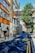 A narrow urban street is lined with brick buildings, one of which has a sign reading 'London Metropolitan University'. The street features parked bicycles, a few pedestrians, and a tree casting shadows across the road. The sky is clear and blue.