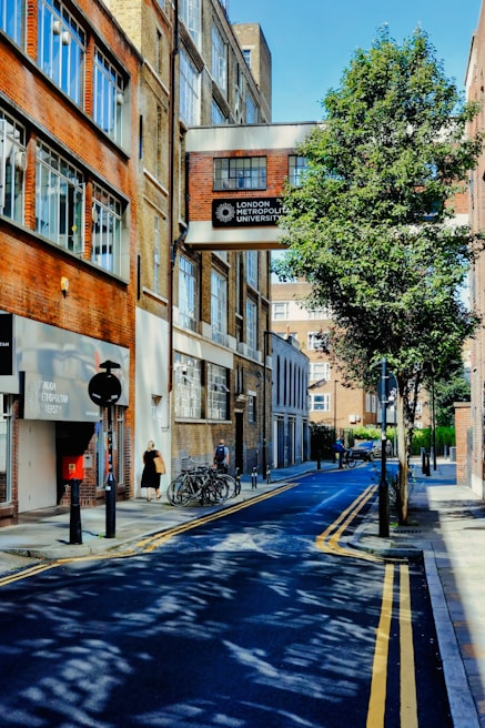 A narrow urban street is lined with brick buildings, one of which has a sign reading 'London Metropolitan University'. The street features parked bicycles, a few pedestrians, and a tree casting shadows across the road. The sky is clear and blue.