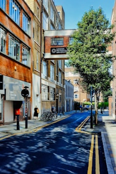A narrow urban street is lined with brick buildings, one of which has a sign reading 'London Metropolitan University'. The street features parked bicycles, a few pedestrians, and a tree casting shadows across the road. The sky is clear and blue.