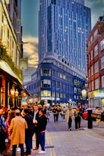A bustling city street scene with people walking and socializing. Tall modern buildings serve as a backdrop, showcasing urban architecture. The sky transitions from day to evening, casting a warm glow on the scene. People are gathering in groups, enjoying conversations and casual strolls.
