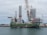 A large vessel docked at a port, equipped with numerous cranes and several large vertical tubular structures, possibly related to wind turbine components. The vessel has a prominent green, red, and white color scheme with the word 'INNOVATION' visible on the side. The surrounding area includes the dock, parts of other machinery, and a cloudy sky.