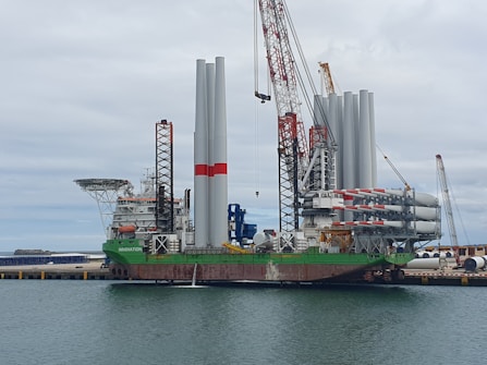 A large vessel docked at a port, equipped with numerous cranes and several large vertical tubular structures, possibly related to wind turbine components. The vessel has a prominent green, red, and white color scheme with the word 'INNOVATION' visible on the side. The surrounding area includes the dock, parts of other machinery, and a cloudy sky.