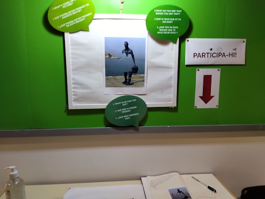 A bulletin board with a central image of a diving statue by the water, surrounded by green speech bubbles that contain questions in multiple languages. A sign on the right reads 'PARTICIPA-HI!' with an arrow below it. A desk below holds an open notebook, a pen, and a bottle of hand sanitizer.