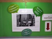 A bulletin board displays a black and white photo of people walking in a city with tall buildings in the background. Surrounding the photo are green speech bubbles with prompts in both English and Spanish. There is a sign that says PARTICIPA with a red arrow pointing downward.