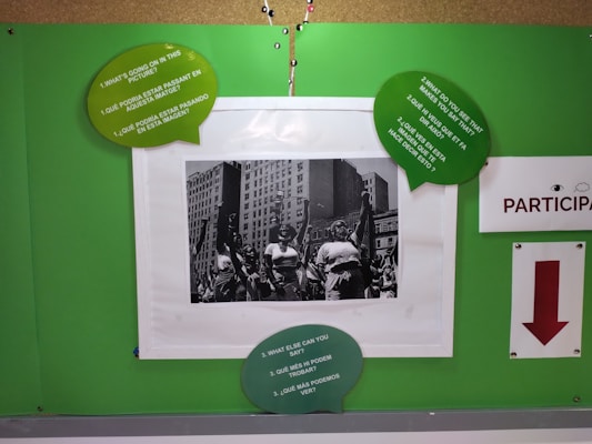 A bulletin board displays a black and white photo of people walking in a city with tall buildings in the background. Surrounding the photo are green speech bubbles with prompts in both English and Spanish. There is a sign that says PARTICIPA with a red arrow pointing downward.