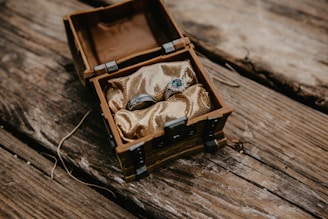An ornate silver ring set with a blue gemstone rests inside a small, rustic wooden chest lined with gold fabric. The chest is situated on a textured wooden surface, suggesting an antique or treasure-like theme.