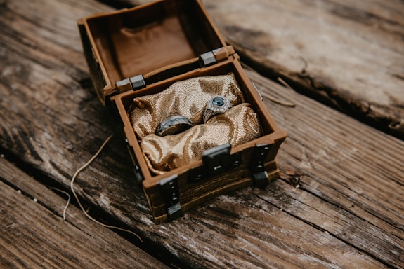 An ornate silver ring set with a blue gemstone rests inside a small, rustic wooden chest lined with gold fabric. The chest is situated on a textured wooden surface, suggesting an antique or treasure-like theme.