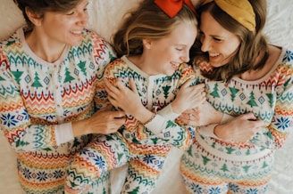 Three people are lying down on a white surface, all dressed in matching colorful patterned pajamas. They appear to be smiling and interacting warmly with each other.
